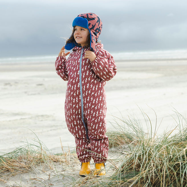 A child on a beach in an EcoSplash waterproof pink puddle suit with lightning bolts, wearing a hat and yellow wellies.